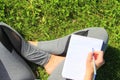 A beautiful young white girl in a white t-shirt and with long hair sitting on green grass, on the lawn and writes with a pen in a Royalty Free Stock Photo