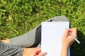 A beautiful young white girl in a white t-shirt and with long hair sitting on green grass, on the lawn and writes with a pen in a Royalty Free Stock Photo