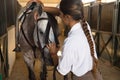 Beautiful young woman adjusting her horse's noseband inside a stable. Concept horse riding, animals, saddle, horsewoman Royalty Free Stock Photo