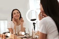 Beautiful young woman applying makeup at table with mirror and ring lamp