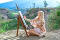 Beautiful young woman artist paints a landscape in nature. Drawing on the easel with colorful paints in the open air. Royalty Free Stock Photo