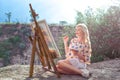 Beautiful young woman artist paints a landscape in nature. Drawing on the easel with colorful paints in the open air. Royalty Free Stock Photo