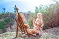 Beautiful young woman artist paints a landscape in nature. Drawing on the easel with colorful paints in the open air. Royalty Free Stock Photo