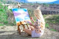 Beautiful young woman artist paints a landscape in nature. Drawing on the easel with colorful paints in the open air. Royalty Free Stock Photo