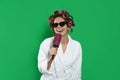 Beautiful young woman in bathrobe with hair curlers and sunglasses singing into hairbrush on green background Royalty Free Stock Photo