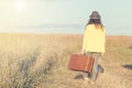 Beautiful young woman with black hat carries brown vintage suitcase in the field road during summer sunset. Back view. Toned image Royalty Free Stock Photo