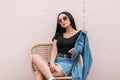 Beautiful young woman in a black top in a denim jacket in sunglasses in a skirt posing on a straw chair near the pink wall Royalty Free Stock Photo