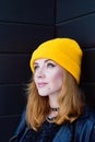 Beautiful young woman with blonde hair and blue eyes in a yellow knitting hat on a background of black wooden wall. Royalty Free Stock Photo