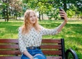 Beautiful young woman with blonde hair taking selfie at the city park. Girl sitting on a bench in a sunny day. Active life concept Royalty Free Stock Photo
