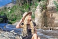 A beautiful young woman blonde with long hair in a hat sitting on a rocky shore by the river. Around the mountains and rocky terra Royalty Free Stock Photo