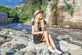A beautiful young woman blonde with long hair in a hat sitting on a rocky shore by the river. Around the mountains and rocky terra Royalty Free Stock Photo