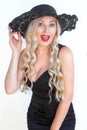 Beautiful young woman blonde with a surprised smile in a black dress and a hat with a decollete. Royalty Free Stock Photo