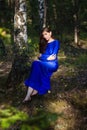 Beautiful, young woman in a blue dress is sitting near a tree over a cliff, against a forest background. Royalty Free Stock Photo