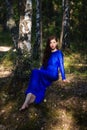 Beautiful, young woman in a blue dress is sitting near a tree over a cliff, against a forest background. Royalty Free Stock Photo