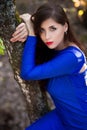 Beautiful, young woman in a blue dress is sitting near a tree over a cliff, against a forest background. Royalty Free Stock Photo