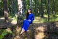 Beautiful, young woman in a blue dress is sitting near a tree over a cliff, against a forest background. Royalty Free Stock Photo