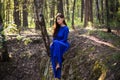 Beautiful, young woman in a blue dress is sitting near a tree over a cliff, against a forest background. Royalty Free Stock Photo