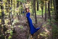 Beautiful, young woman in a blue dress stands near a tree over a cliff, against a forest background. Royalty Free Stock Photo