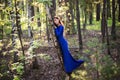 Beautiful, young woman in a blue dress stands near a tree over a cliff, against a forest background. Royalty Free Stock Photo