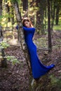 Beautiful, young woman in a blue dress stands near a tree over a cliff, against a forest background. Royalty Free Stock Photo