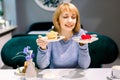 Beautiful young woman in blue sweater is having tea and cake in a nice cafe. Woman choosing between red and yellow tasty Royalty Free Stock Photo