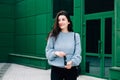 Beautiful young woman in a blue wool sweater posing in city street on green building urban background. Confident business lady,