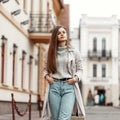 Beautiful young woman with brown hair in a stylish long trench coat in a knitted sweater in a blue jeans walks on the city.