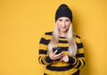 Beautiful young woman browse on smartphone, model wearing woolen cap and sweater, isolated on yellow background. Trendy girl