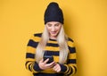 Beautiful young woman browse on smartphone, model wearing woolen cap and sweater, on yellow background. Trendy girl