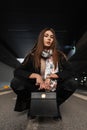 Beautiful young woman brunette in stylish black elegance clothes with fashionable leather handbag is sitting on the asphalt in the Royalty Free Stock Photo