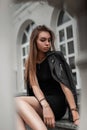 Beautiful young woman with chic brown long hair in a black stylish dress in a vintage black leather jacket posing near a white Royalty Free Stock Photo