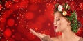 Beautiful young woman with Christmas wreath blowing magical snowy dust on red background