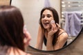 Beautiful young woman is cleaning her face using a cotton disc and smiling while looking in the mirror in the bathroom Royalty Free Stock Photo