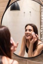 Beautiful young woman is cleaning her face using a cotton disc and smiling while looking in the mirror in the bathroom Royalty Free Stock Photo