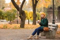 Beautiful young woman with coffee resting on wooden bench in autumn park Royalty Free Stock Photo