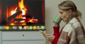 Beautiful young woman in cozy warm sweater with cup of hot drink on the background of the fireplace and Christmas lights Royalty Free Stock Photo