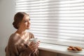 Beautiful young woman with cup of hot drink and cookies near window at home Royalty Free Stock Photo