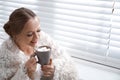 Beautiful young woman with cup of hot drink and marshmallow near window at home Royalty Free Stock Photo