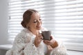 Beautiful young woman with cup of hot drink near window. Winter atmosphere Royalty Free Stock Photo