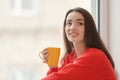 Beautiful young woman with cup of hot tea near window at home Royalty Free Stock Photo