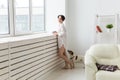 Calm and coziness. Beautiful young woman with cup of tea and dog standing near the window at home Royalty Free Stock Photo