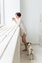 Calm and coziness. Beautiful young woman with cup of tea and dog standing near the window at home Royalty Free Stock Photo