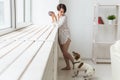 Calm and coziness. Beautiful young woman with cup of tea and dog standing near the window at home Royalty Free Stock Photo