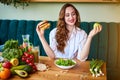 Beautiful young woman decides eating hamburger or apple in kitchen. Cheap junk food vs healthy diet Royalty Free Stock Photo