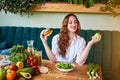 Beautiful young woman decides eating hamburger or apple in kitchen. Cheap junk food vs healthy diet Royalty Free Stock Photo