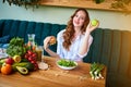 Beautiful young woman decides eating hamburger or apple in kitchen. Cheap junk food vs healthy diet