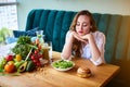 Beautiful young woman decides eating hamburger or vegetables in kitchen. Cheap junk food vs healthy diet