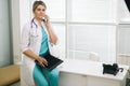 Beautiful young woman doctor with clipboard standing and talking on cell phone Royalty Free Stock Photo