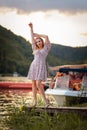 A beautiful young woman in a dress poses standing on a river pier. Vertical. A mountain and boats in the background Royalty Free Stock Photo