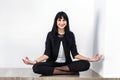 Beautiful young woman dressed in a black business suit sitting in lotus position on a floor in office, smiling, looking at camera